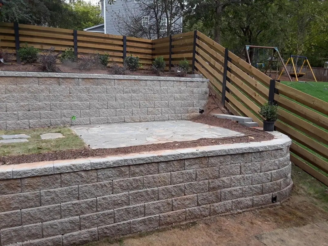 Horizontal board fence with black posts above a stone retaining wall and patio